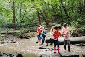 Group of school children and their teachers working together to cross a river. The older students are giving a helping hand to the younger students to cross and stand on the wet rocks.